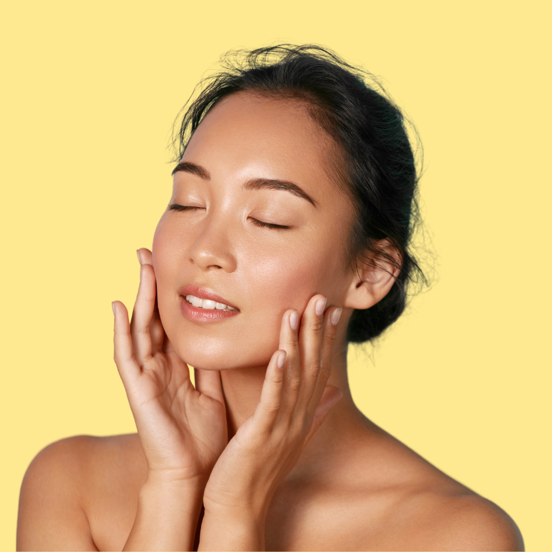 close-up of a woman with dewy, glowing skin, eyes closed, hands resting on her cheeks on a soft yellow background