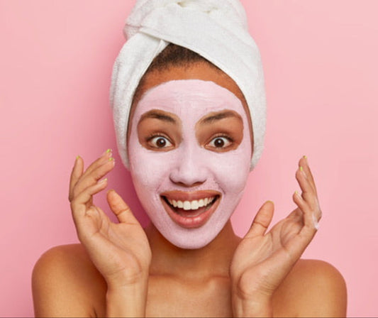 Smiling model with Moirai Berry and Bright face mask applied, towel wrapped hair on a pink background.