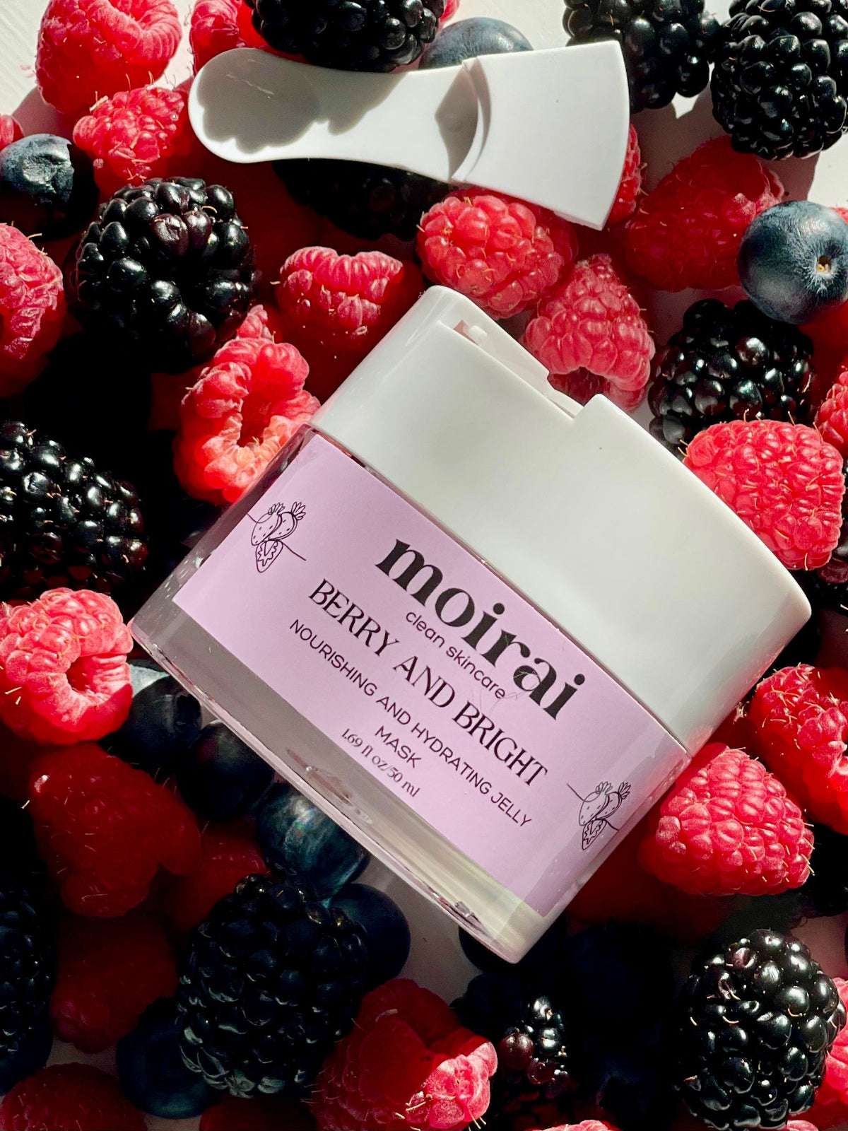 A jar of 'Berry and Bright Face Mask' by Moirai Skincare placed among an array of fresh berries, including raspberries, blueberries, and blackberries.