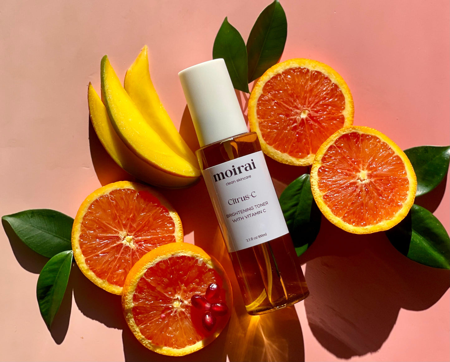 Moirai Citrus-C Brightening Toner styled with pink grapefruit slices, mango and green leaves on a pink background.