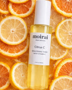 Moirai Citrus C Brightening Toner with Vitamin C in a clear yellow bottle surrounded by fresh orange and lemon slices
