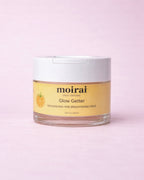 Moirai Glow Getter Nourishing and Brightening Moirai Glow Getter Nourishing and Brightening Mask in a clear jar with white lid on a soft blush pink background.