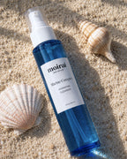 A blue glass bottle of Moirai Marine Complex Hydrating Cleanser placed on sandy ground with seashells around it.