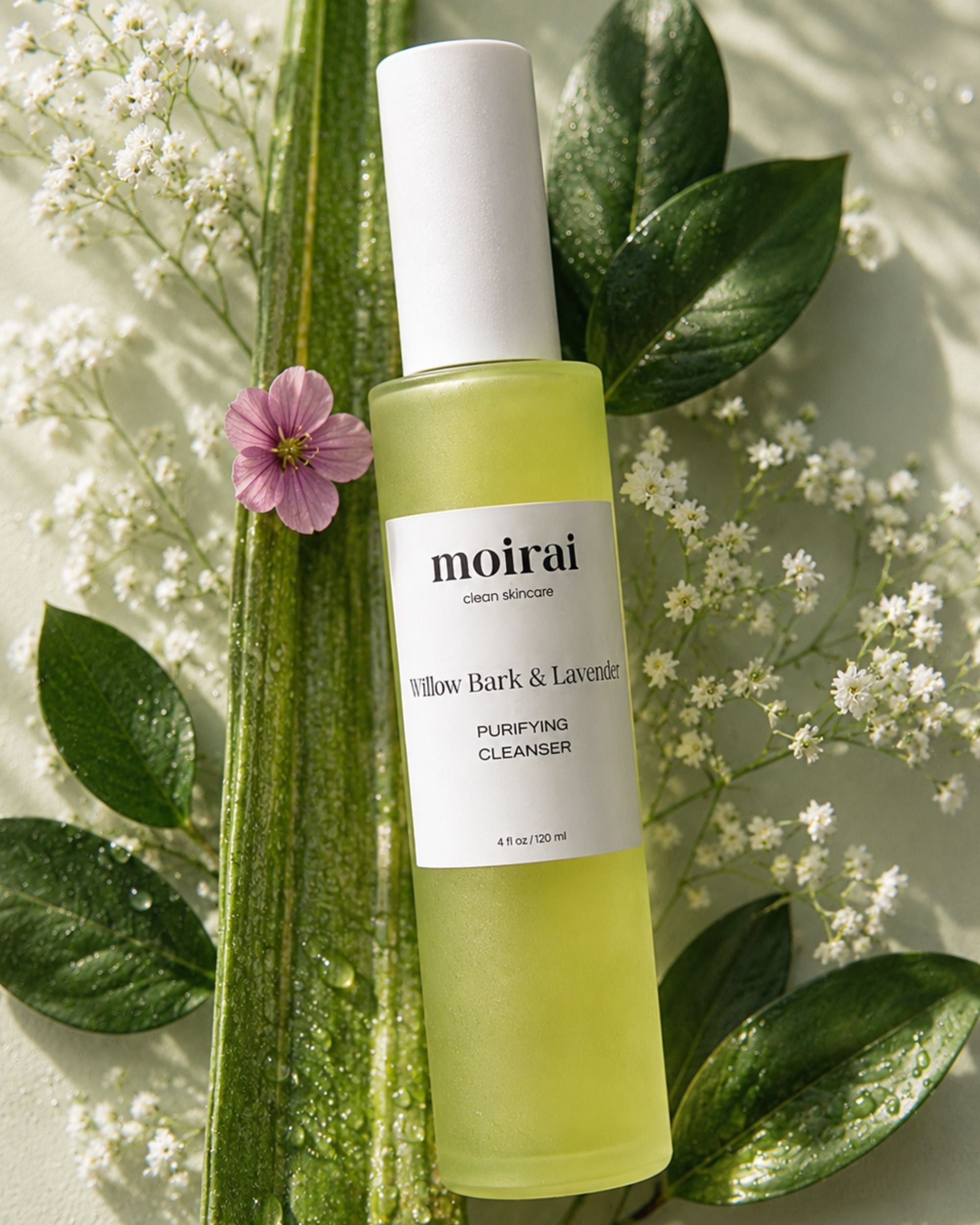 A bottle of Moirai Willow Bark & Lavender Purifying Gel Cleanser styled with green leaves and white flowers on a light background.