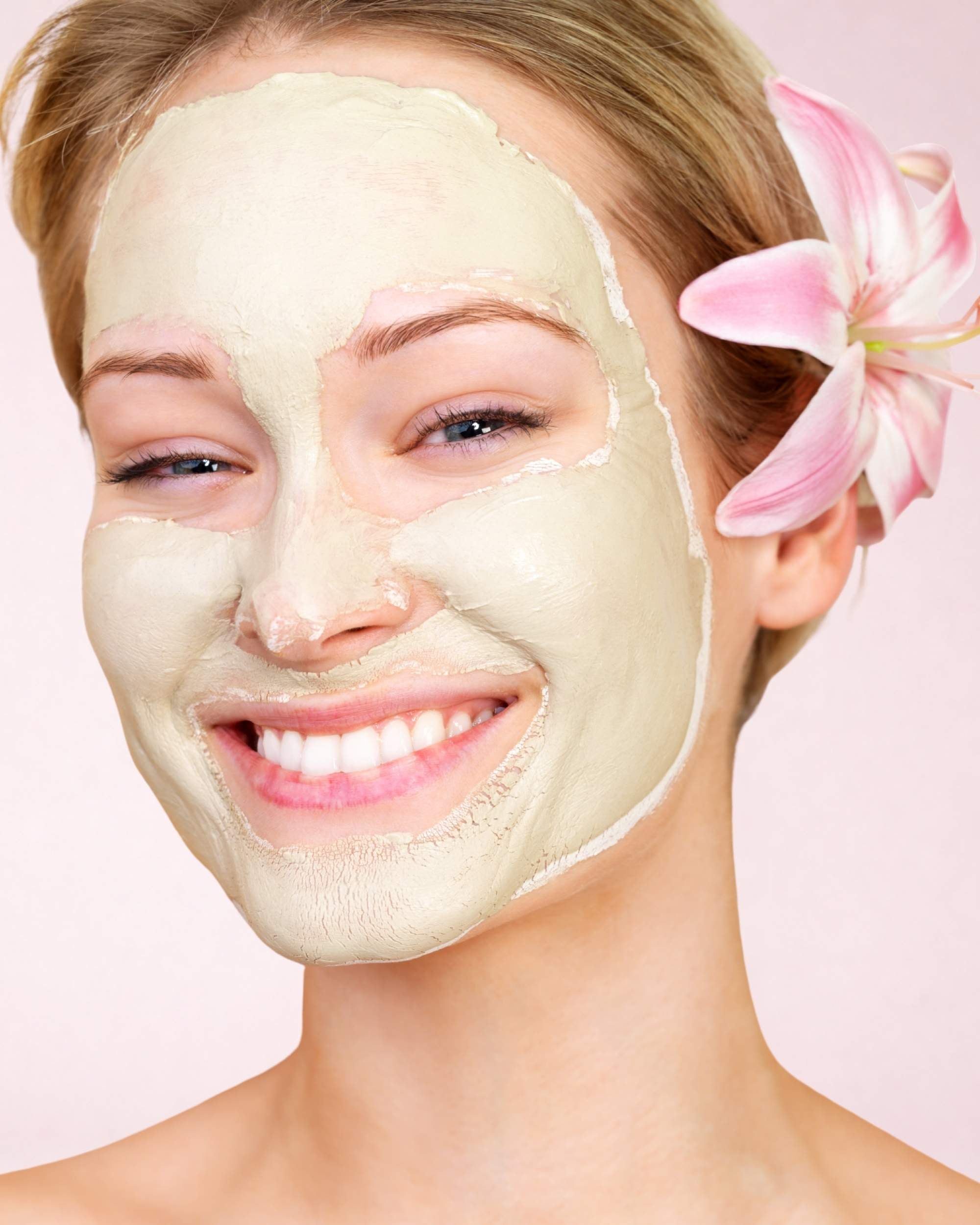 Woman wearing a facial mask with a pink flower in her hair on a soft blush background.